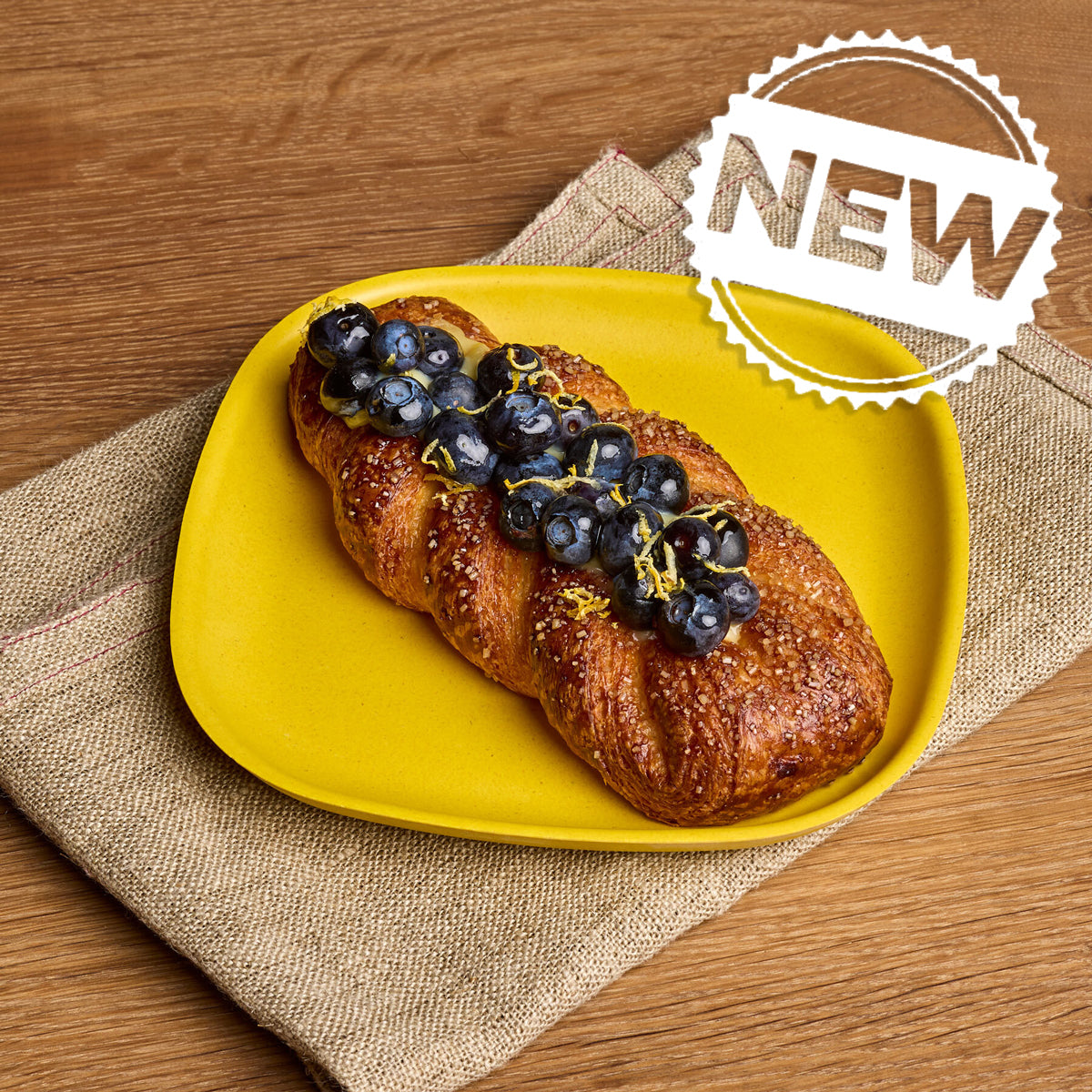 A braided pastry topped with blueberries and a thin yellow zest strip, served on a bright yellow plate over a beige napkin on a wooden table. A white 'NEW' badge is in the top right corner.