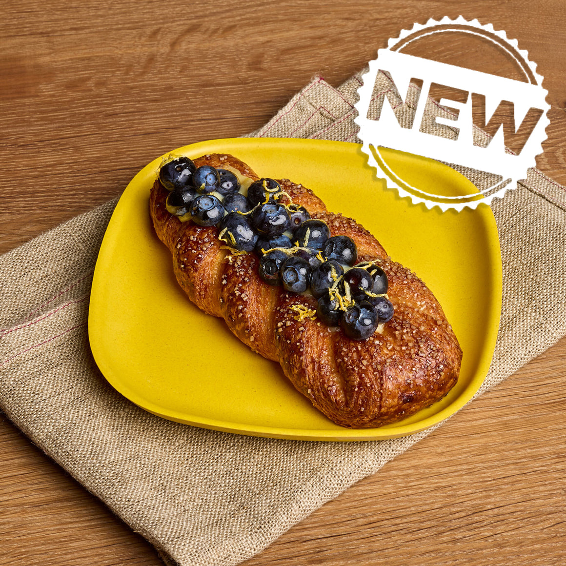 A braided pastry topped with blueberries and a thin yellow zest strip, served on a bright yellow plate over a beige napkin on a wooden table. A white 'NEW' badge is in the top right corner.