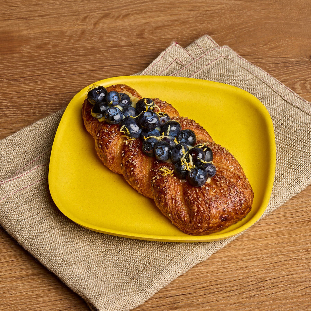 A braided croissant topped with fresh blueberries and a drizzle of yellow zest, served on a bright yellow plate over a beige woven placemat on a wooden table.