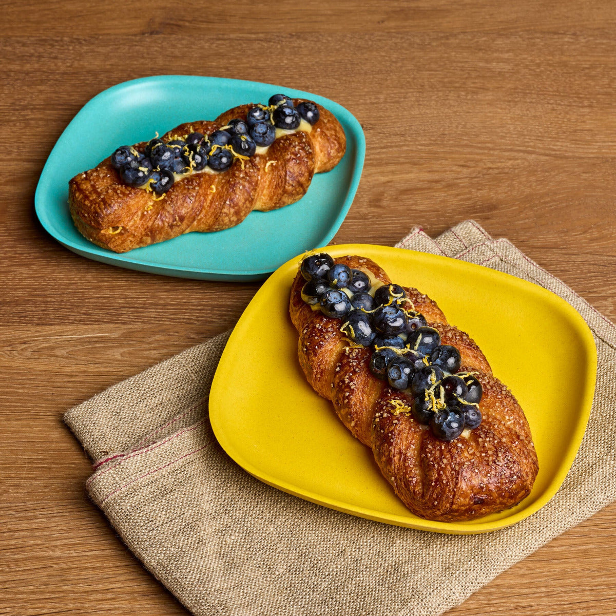 Two braided pastries topped with blueberries and yellow zest, served on colorful plates on a wooden table with a burlap napkin. One pastry is on a turquoise plate and the other is on a yellow plate.