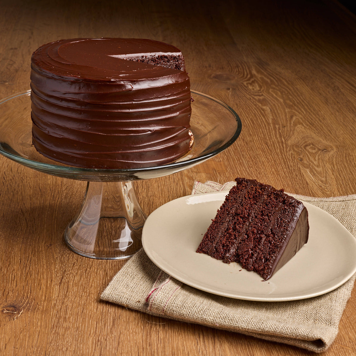 A chocolate cake with smooth dark chocolate frosting sits on a glass pedestal plate, with a slice placed on a beige plate on a wooden table.