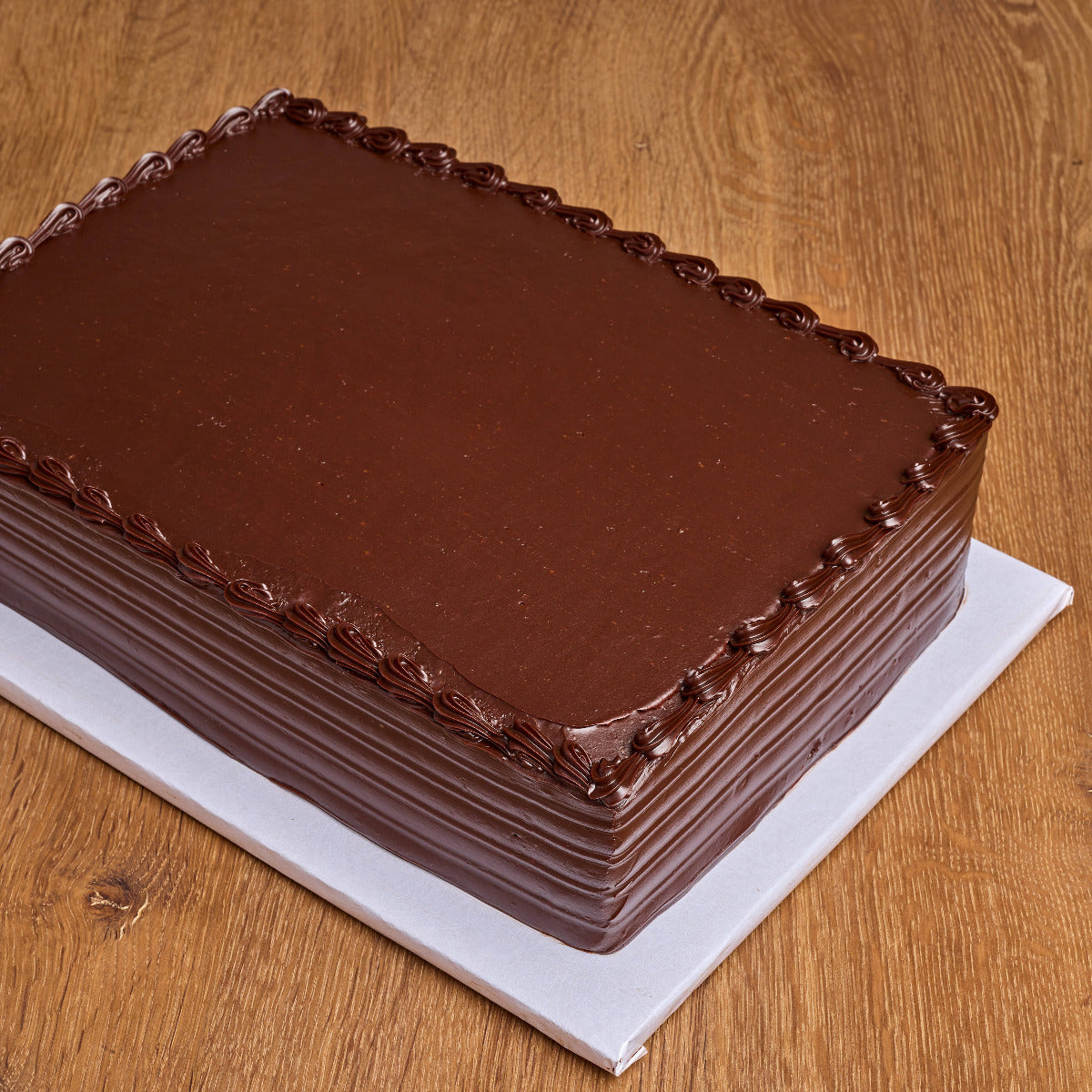 A rectangular chocolate cake with smooth dark chocolate frosting and piped decorative borders on top, presented on a white board over a wooden surface.